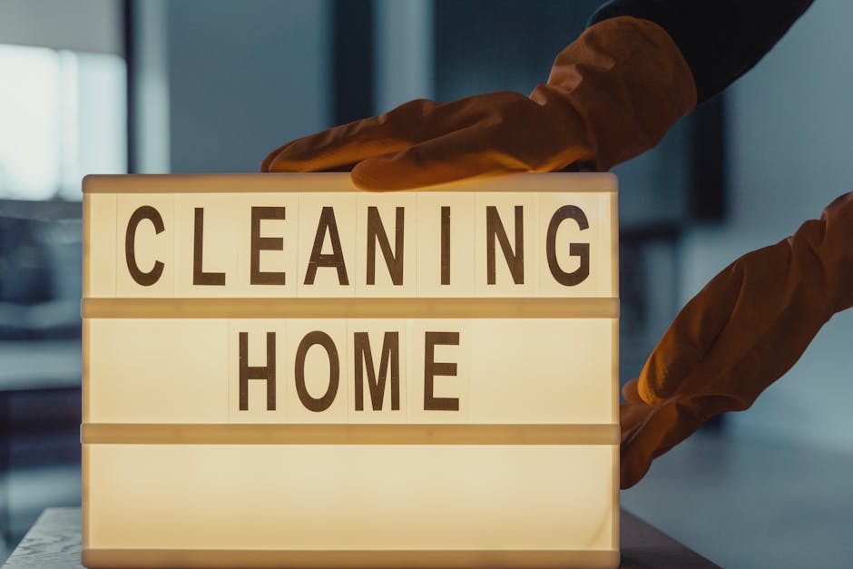 A person wearing orange rubber gloves is adjusting or placing a lightbox sign with black letters spelling 'CLEANING HOME' on a wooden surface. The background features a modern, softly lit interior wit